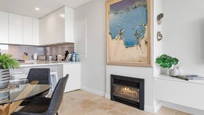 Una cocina moderna con armarios blancos, una zona de comedor con una mesa y sillas de vidrio, una chimenea y un cuadro con temática playera en la pared.