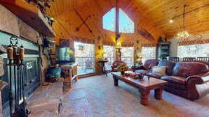 L’intérieur d’un chalet rustique, avec une cheminée en pierre, un canapé en cuir et une grande fenêtre offrant une vue sur l’extérieur.