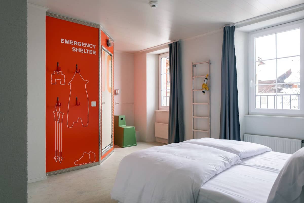 A hotel room with a bed, a ladder, a green chair, and a window with curtains.