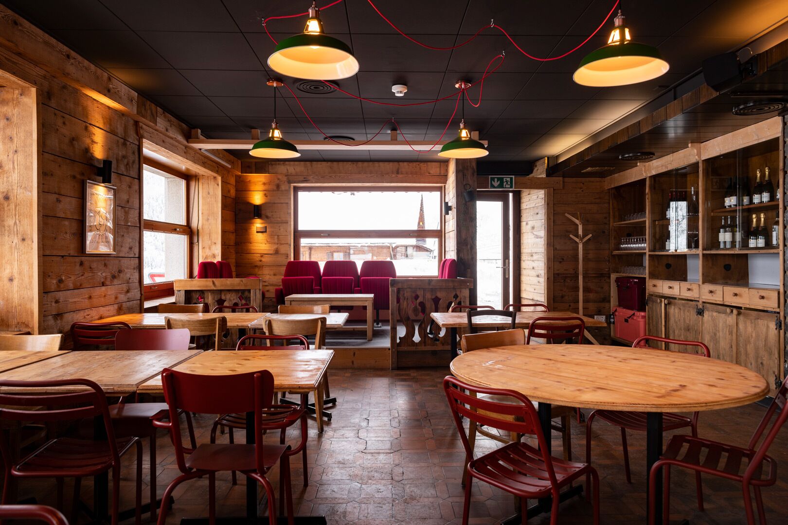 A cozy restaurant with wooden walls and tables, red chairs, and hanging pendant lights.