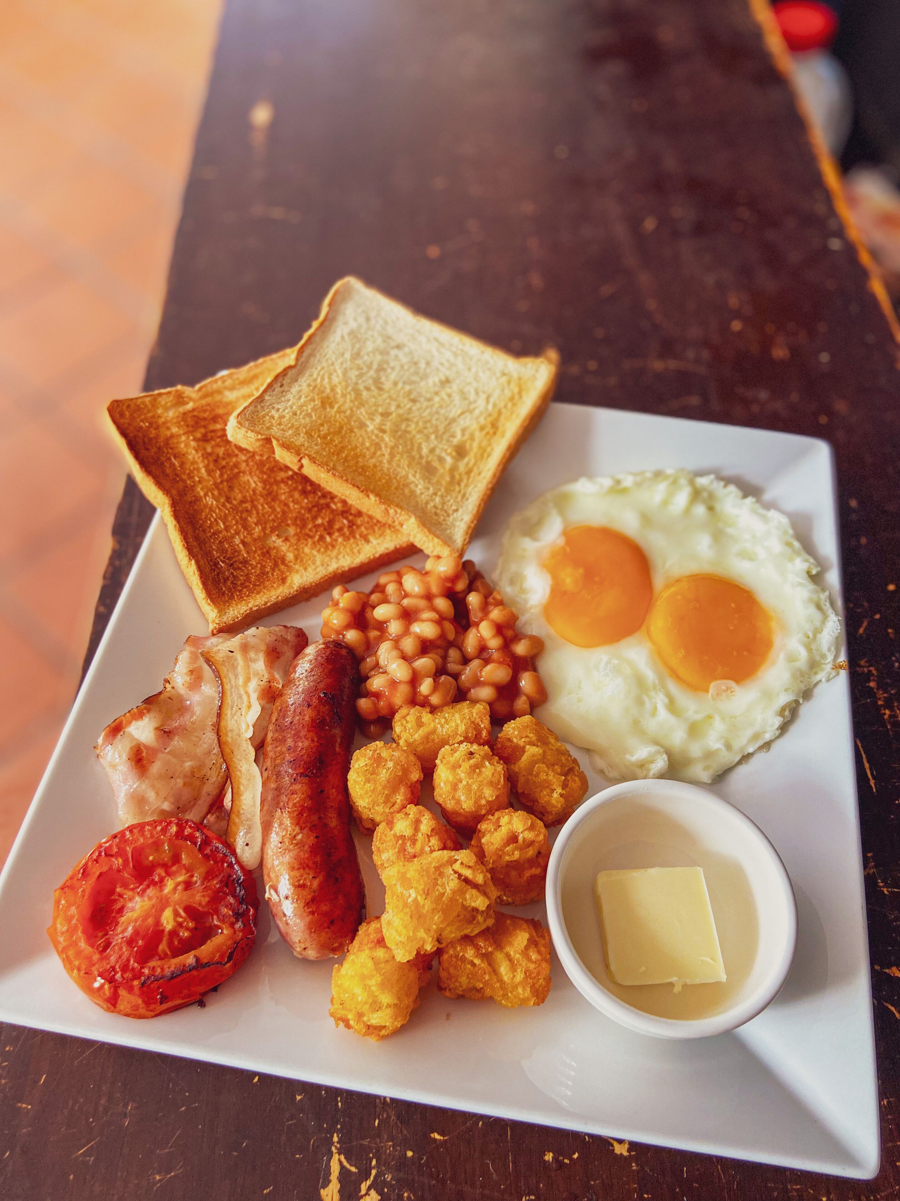 A breakfast plate with toast, eggs, beans, sausages, fried tomatoes, and a small bowl of butter.