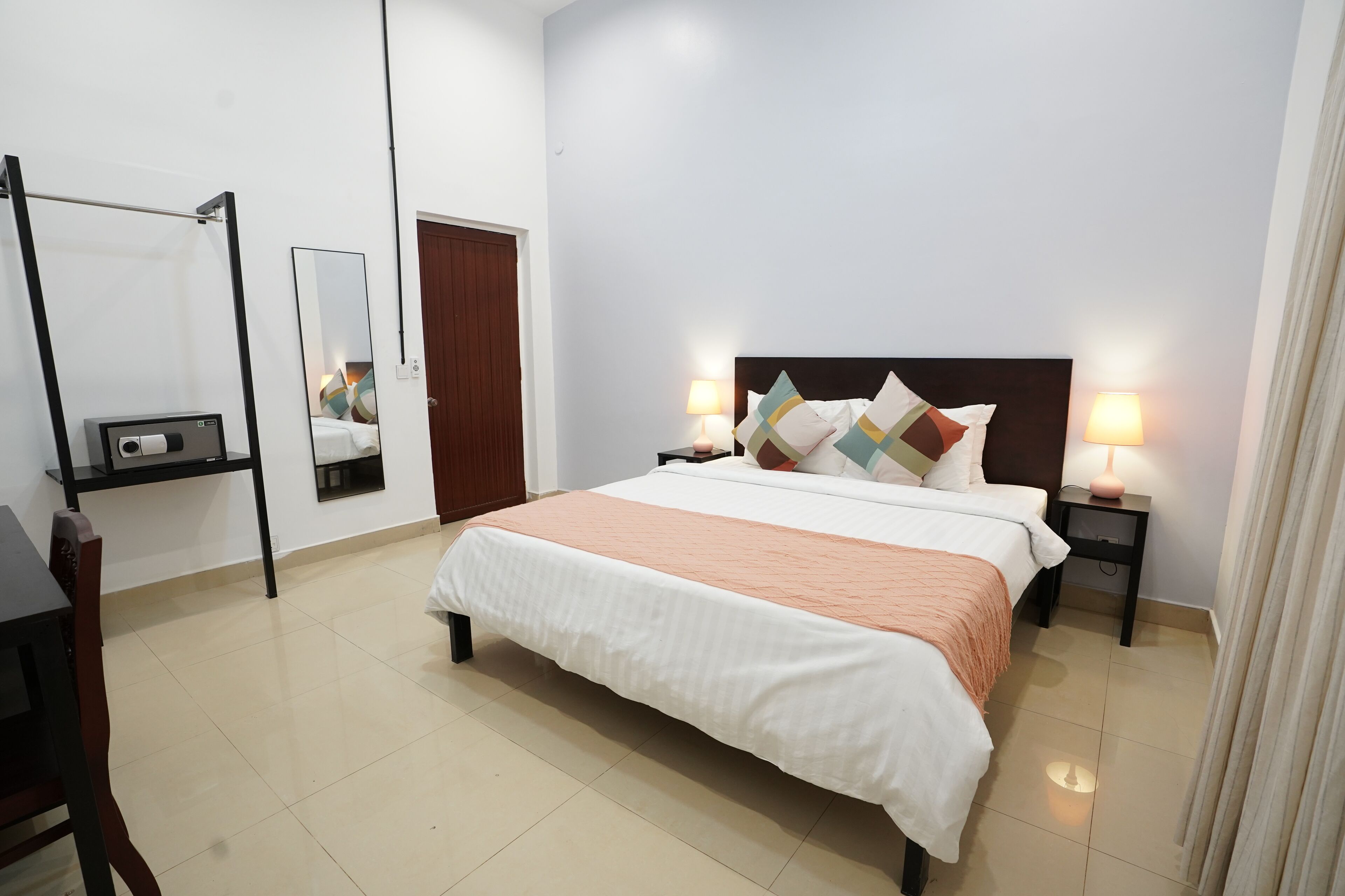 A hotel room with a large bed, two bedside tables with lamps, a mirror, a door, and a small table with a chair.