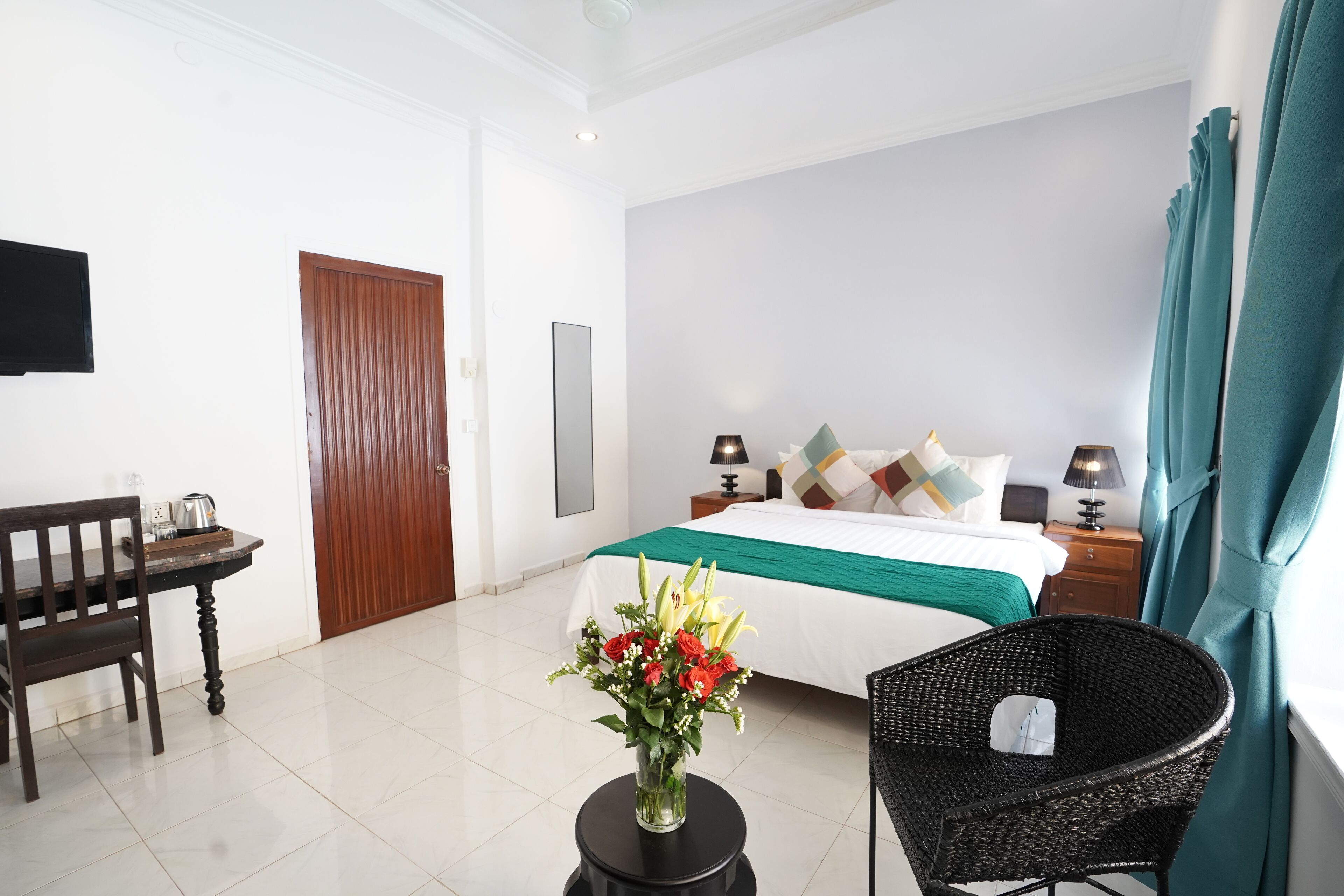 A hotel room with a bed, a chair, a small table with a vase of flowers, a wooden desk, and a television on the wall.