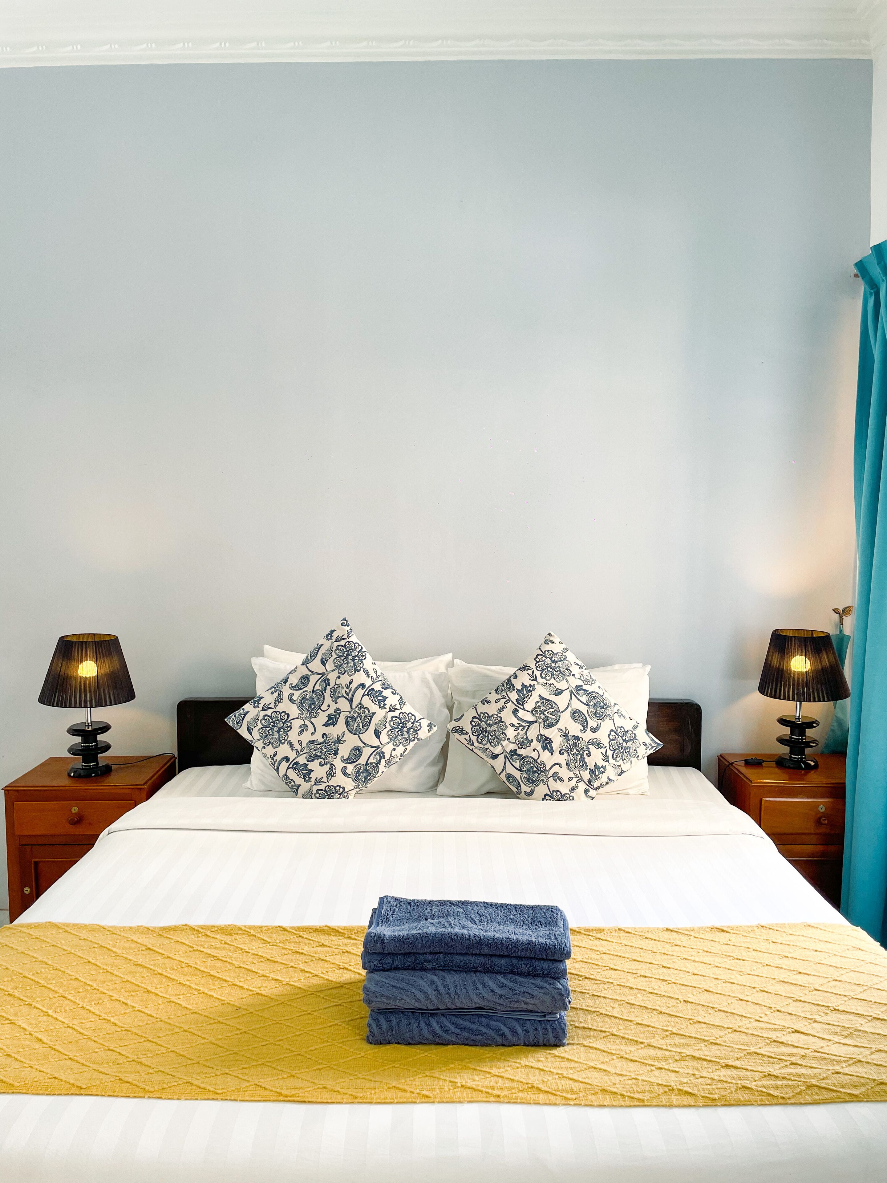 A neatly made bed with white bedding and blue floral pillows, two bedside lamps, and a folded blue towel on a yellow rug.