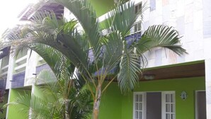A two-story building with a green facade, a tiled roof, and a small garden with palm trees and red flowers. - Village Duplex, 2 Bedrooms, 100m from the Beach with Pool (Salvador)