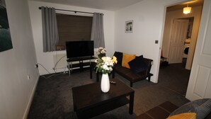 Ein Wohnzimmer mit einem dunkelbraunen Sofa, einer weißen Vase mit Blumen, einem Flachbildfernseher und einem Fenster mit grauen Vorhängen.