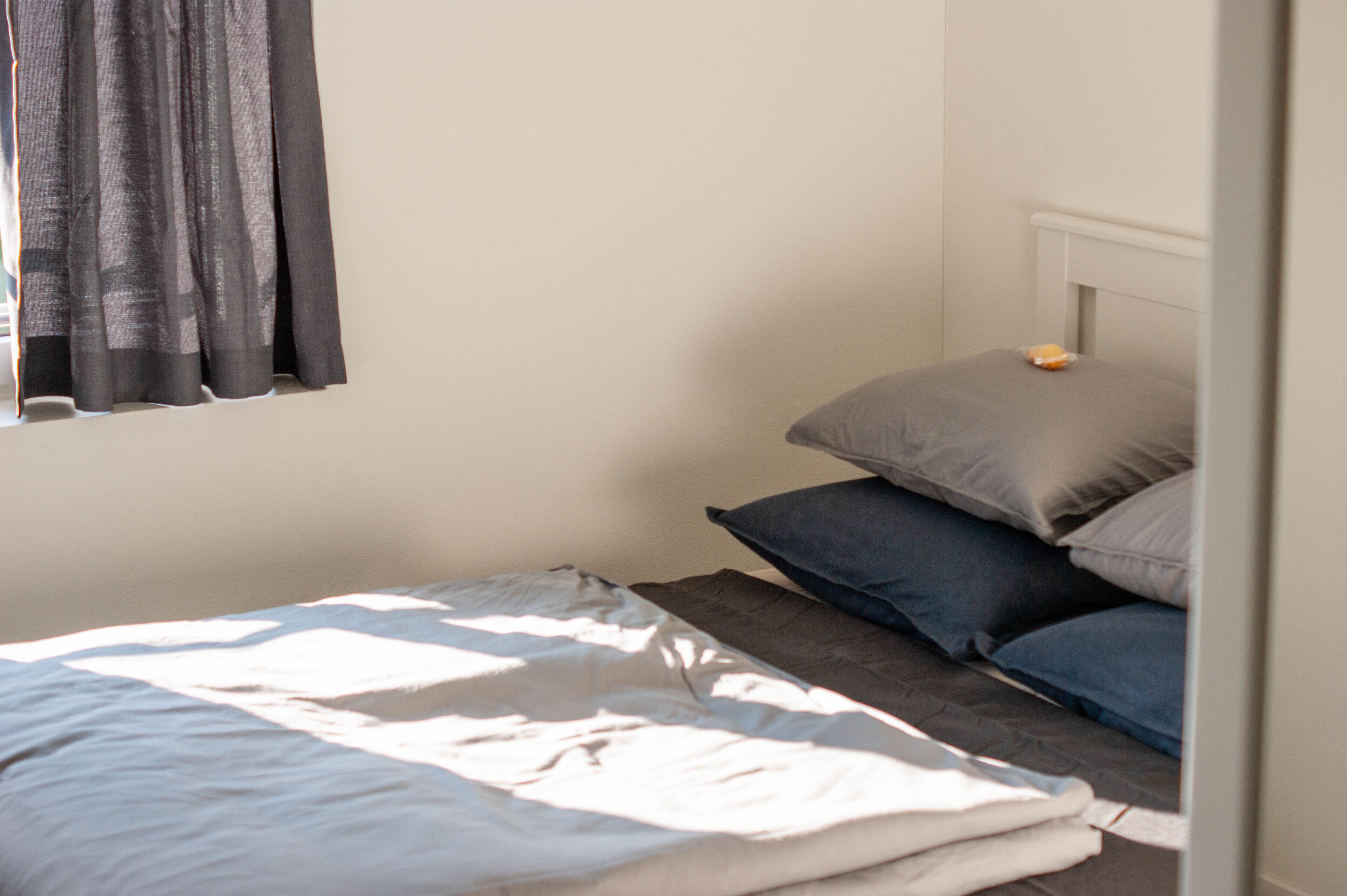 Una cama individual con edredón blanco y gris, una ventana con cortinas grises y un cabecero blanco.