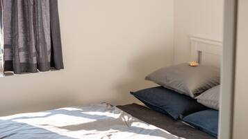 Una cama individual con edredón blanco y gris, una ventana con cortinas grises y un cabecero blanco.