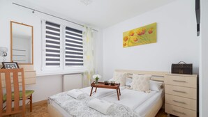 A bedroom with a bed, a wooden chair, a dresser, a bedside table with a tray, a window with blinds, and a painting on the wall.