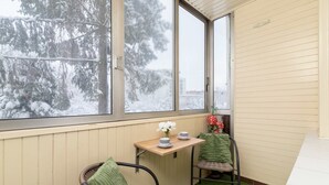 A cozy corner with a window, a small table with a vase and cups, a chair with a green towel, and a view of snow-covered trees.