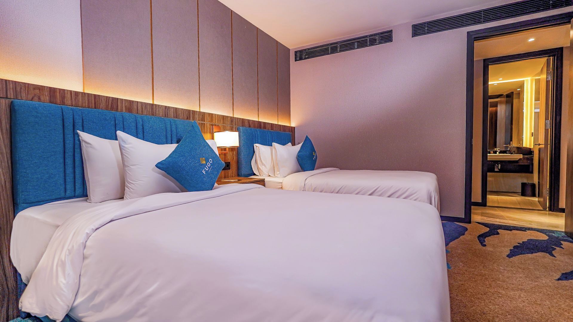 Een hotelkamer met twee bedden, een houten hoofdbord, een nachtkastje, een lamp en een badkamer die door een geopende deur zichtbaar is.