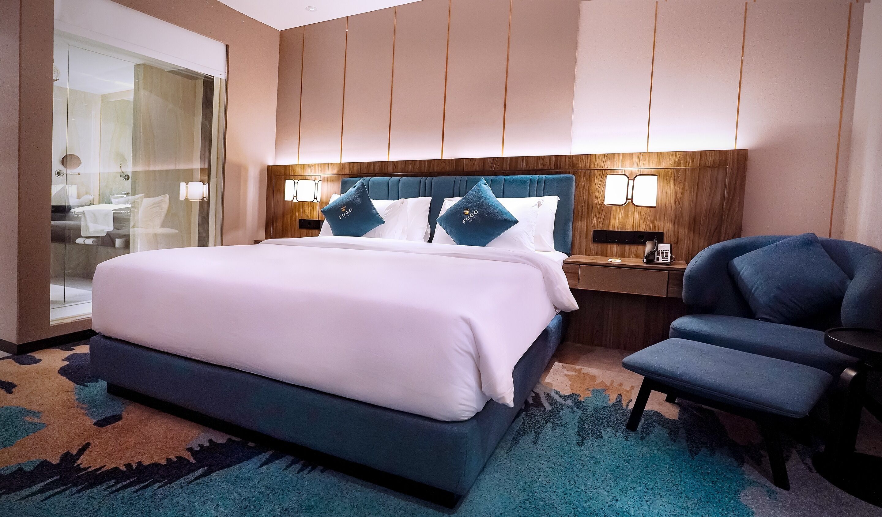 Ein Hotelzimmer mit einem großen Bett, einem Nachttisch mit Telefon, einem Sessel und einer Duschkabine mit Glas.