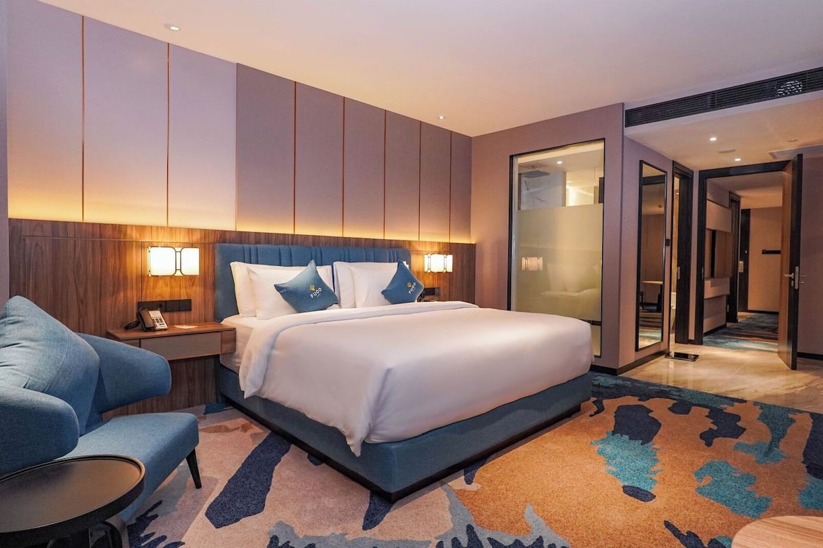 Ein modernes Hotelzimmer mit einem großen Bett, einer Sitzecke mit einem Sessel und einem kleinen Tisch sowie einer Glastür zu einem Flur.