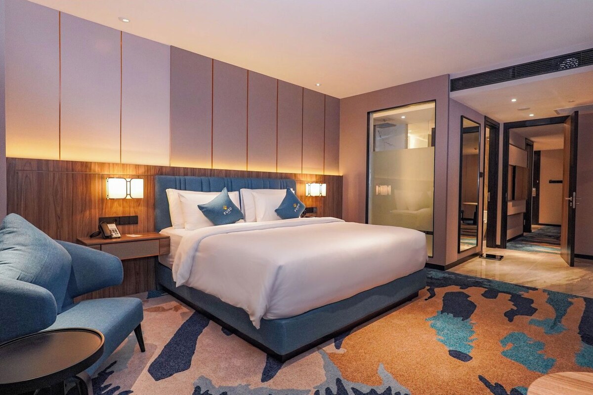 Una habitación de hotel moderna con una cama grande, un área de descanso con una silla y una mesita, y una puerta de vidrio que da a un pasillo.