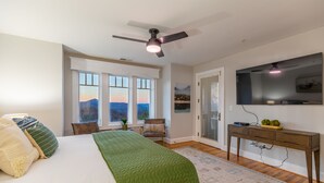 Un dormitorio con una cama, un edredón verde, un escritorio con televisor y vistas a la montaña desde la ventana.
