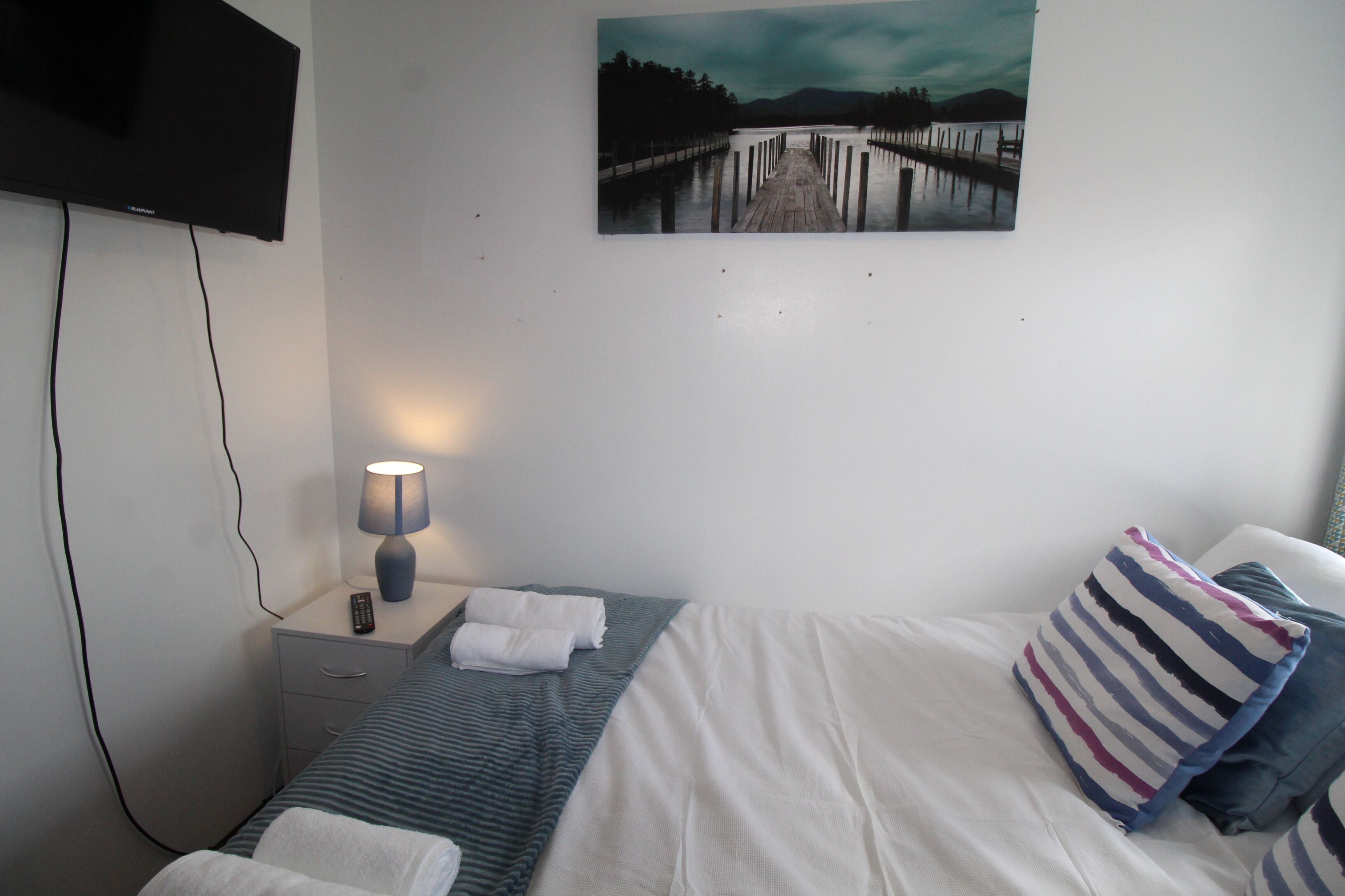 Quarto de hotel com cama, mesa de cabeceira com abajur, controle remoto e um quadro na parede.