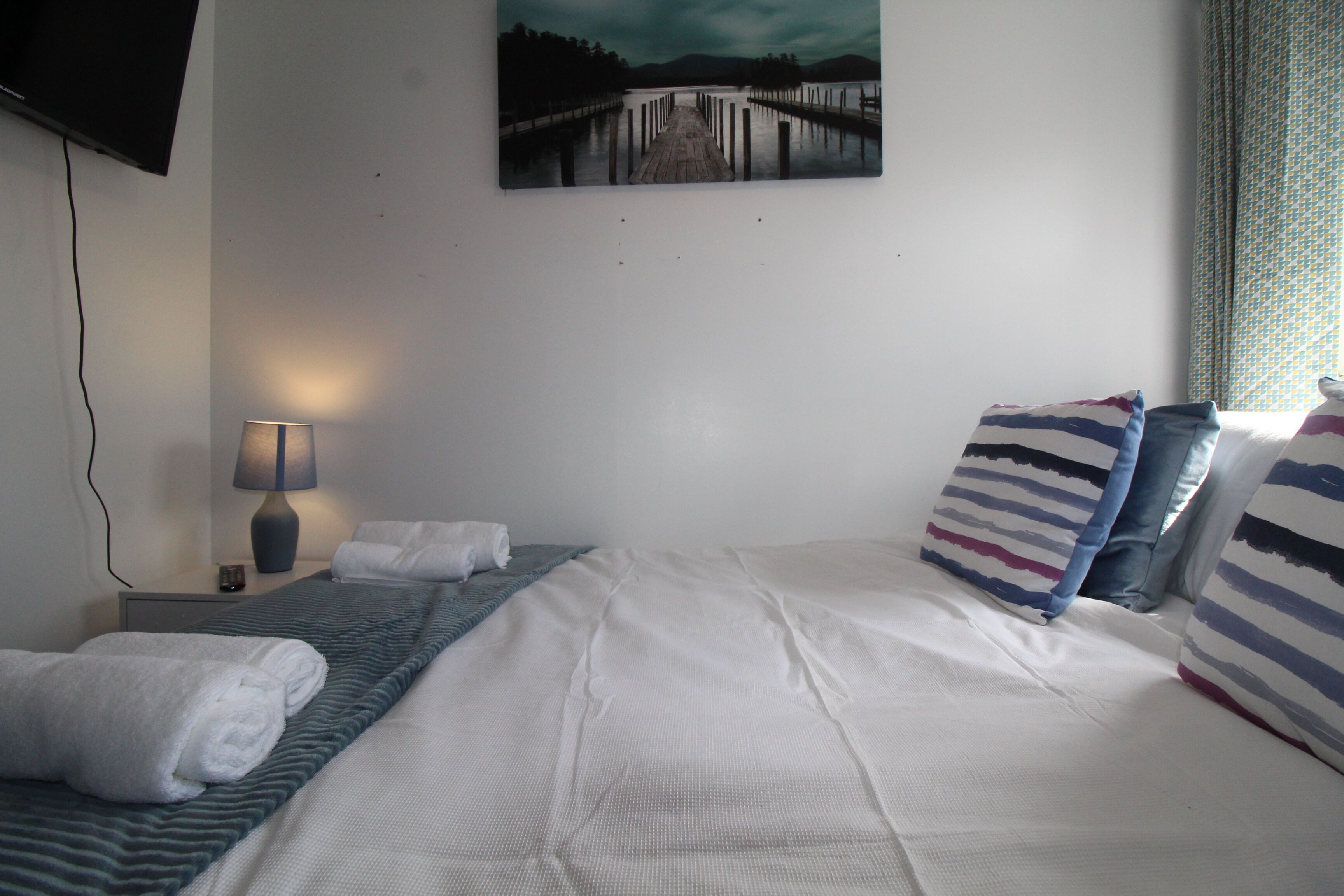 A neatly made bed with white linens and striped pillows, a bedside lamp, and a painting of a pier on the wall.
