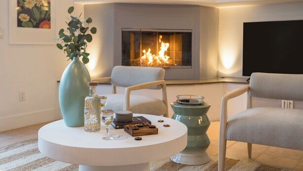 A modern living room with a fireplace, a round coffee table with a vase and a bottle, and two chairs.