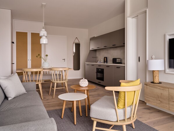 A modern living room with a grey sofa, wooden chairs, a small round table, and a kitchen area with cabinets and appliances. - the breeze (Heringsdorf)