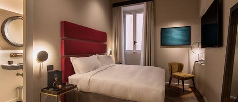 A modern bedroom with a large bed, a red accent wall, a bedside table with a lamp, and a television mounted on the wall.