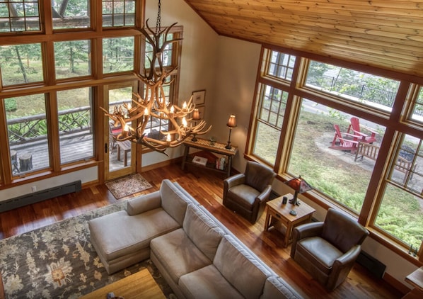 A spacious living room with a large chandelier, leather sofas, and a view of a deck with outdoor furniture. - Gorgeous lake views, walk to village, luxury chalet with 6,000+ sq ft (Lake Placid)
