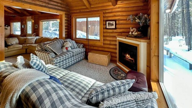 L’intérieur chaleureux d’un chalet avec une cheminée, une literie à carreaux et une vue sur une forêt enneigée.