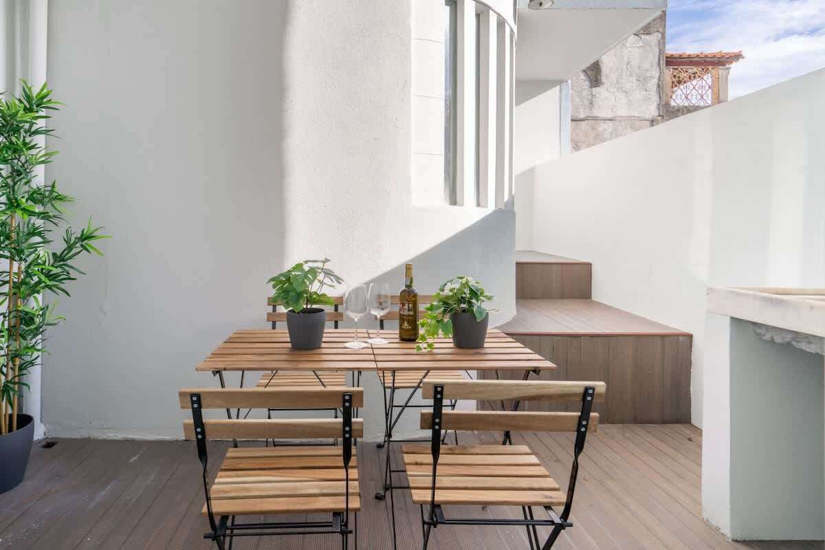 Uma mesa de madeira com cadeiras, plantas em vasos e uma garrafa de vinho numa varanda com parede branca e escada.