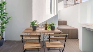 Uma mesa de madeira com cadeiras, plantas em vasos e uma garrafa de vinho numa varanda com parede branca e escada.