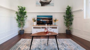 Quarto com uma mesa de madeira, duas plantas em vasos, uma televisão exibindo 'hostwise' e um quadro de um barco na parede.