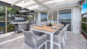 Eine Außenpatio mit Holztisch, Korbstühlen, einem Grill und einer Glastür, die zu einem überdachten Bereich führt.