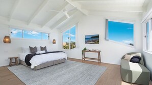 Een ruime slaapkamer met een groot bed, een houten nachtkastje, een klein tafeltje en uitzicht op zee door de ramen.