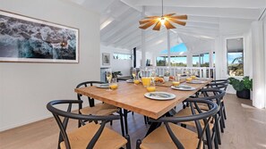 Un coin repas avec une table en bois dressée pour six personnes, entourée de chaises noires à assise tissée. Un grand tableau représentant un paysage naturel est accroché au mur.