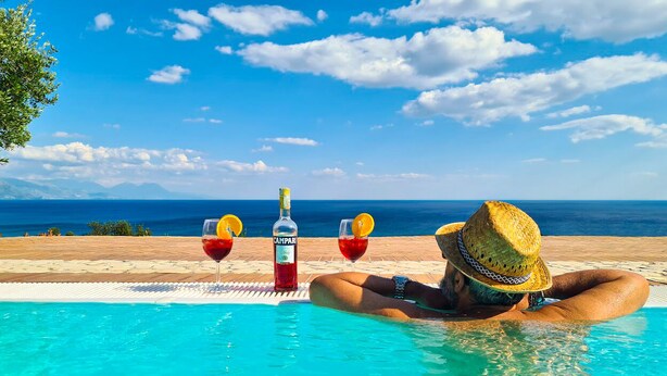 Una persona relajándose en una piscina con una botella de Campari y dos vasos de vino tinto.