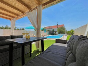 Een terras buiten met een zwembad, een overdekte zithoek en een huis op de achtergrond.