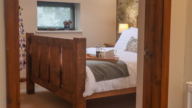 A bedroom with a wooden bed, a window with a view, a bedside table with a lamp, and a tray with items. - The Coach House (Cheltenham)