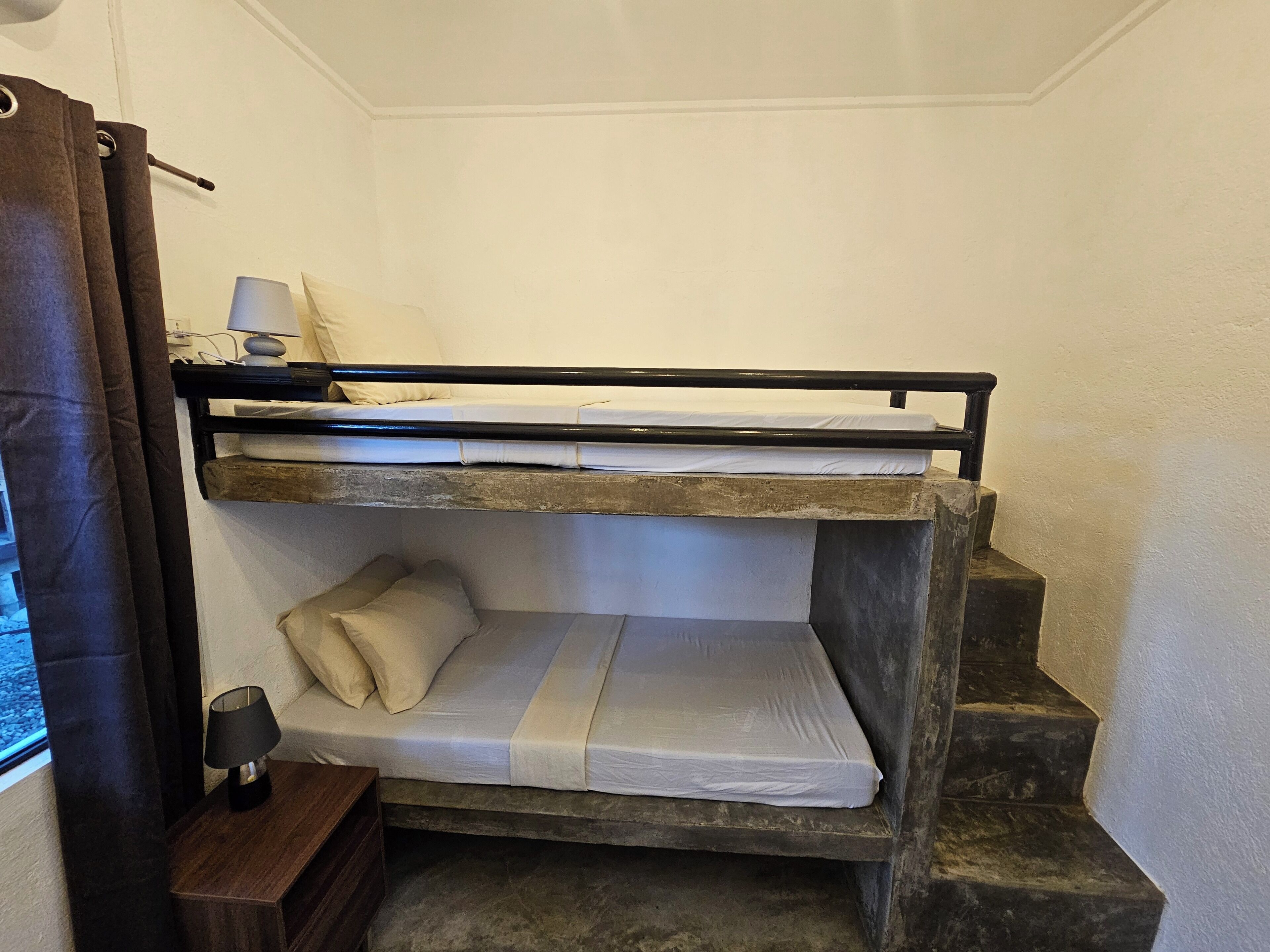 Een stapelbed met houten frame, een klein nachtkastje met lamp, en een raam met gordijnen.