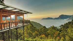 Una estructura de madera con cerramiento de vidrio, elevada sobre pilotes, con vistas a un valle exuberante y montañas distantes, además de un mar de nubes.