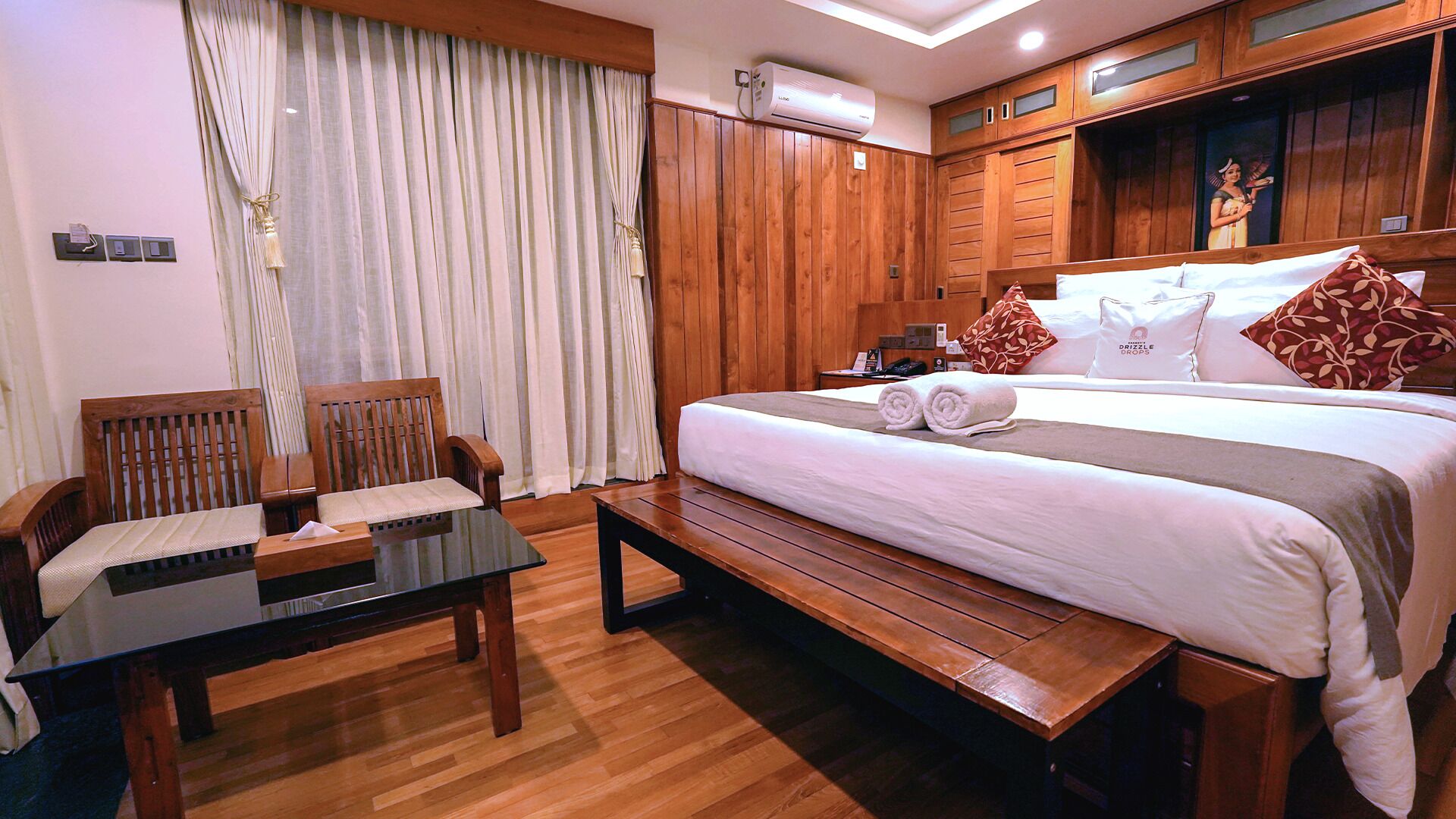 A hotel room with a wooden floor, a bed with white bedding and two rolled towels, a wooden bench, a small table, and a wooden wardrobe.