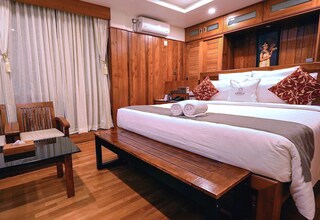 A hotel room with a wooden floor, a bed with white bedding and two rolled towels, a wooden bench, a small table, and a wooden wardrobe.