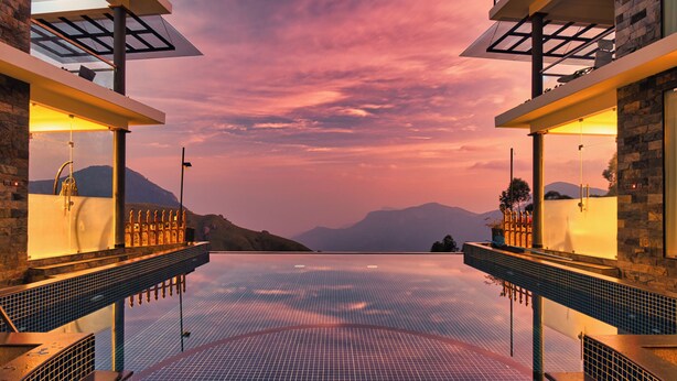 Una piscina infinita moderna con vistas a montañas al atardecer, rodeada de muros de piedra y barandas de vidrio.