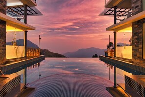 Una piscina infinita moderna con vistas a montañas al atardecer, rodeada de muros de piedra y barandas de vidrio.