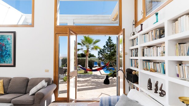 A spacious living room with a large window, a hammock, a bookshelf, and a pool view.