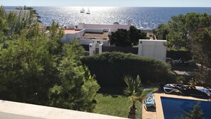A coastal view with a swimming pool, a building with a tiled roof, and sailboats on the sea.
