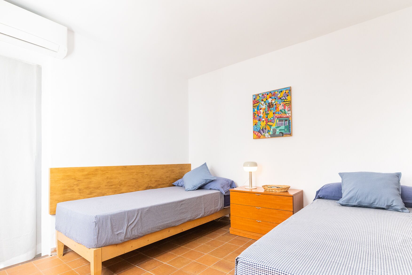 A bedroom with two single beds, a wooden headboard, a nightstand with a lamp, and a colorful artwork on the wall.