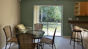 A dining area with a glass table and wicker chairs, a ceiling fan, and a sliding glass door leading to a garden. - Gemme au paradis (Lake Worth)