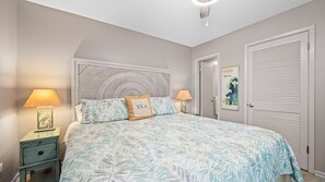 Une chambre à coucher avec un lit, deux tables de chevet avec des lampes, une tête de lit, une porte ornée d’une affiche et une armoire à portes à persiennes.