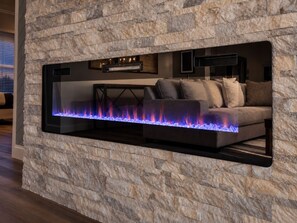 Una sala moderna con pared de textura de piedra, chimenea empotrada con llama azul, sofá oscuro y televisor de pantalla plana sobre la chimenea.