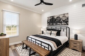 Un dormitorio con cama, mesitas de noche, un banco, ventilador de techo, ventana con persianas y una obra de arte colgada en la pared.
