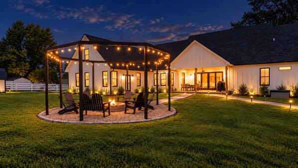 A covered outdoor seating area with chairs and a fire pit, surrounded by a well-maintained lawn.