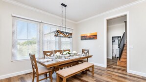 Un comedor con una mesa de madera, sillas y un banco. Se observan paredes blancas, un cuadro y una escalera al fondo.